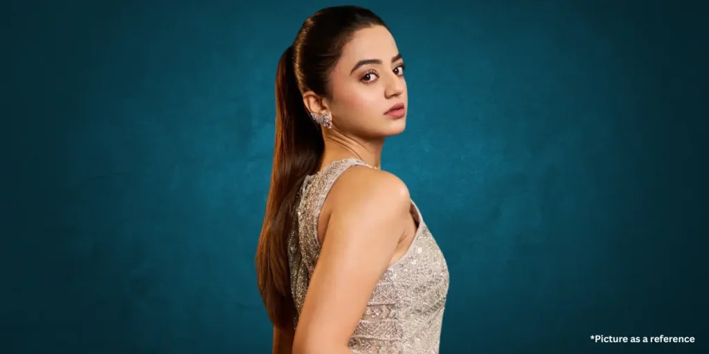 A confident actress Helly Shah dressed in a glittering silver sleeveless gown with a sleek high ponytail hairstyle. She poses elegantly against a rich, textured teal background.