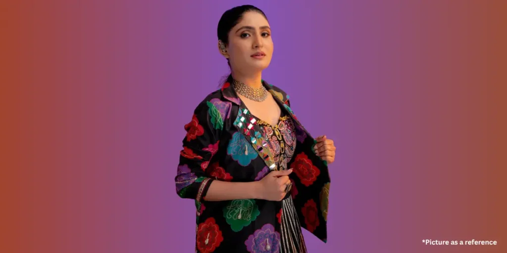 A confident woman, Geetaben Rabari, wearing a modern black jacket with multicolored floral patterns and mirror embellishments, paired with traditional jewelry, standing against a trendy purple and orange gradient background.