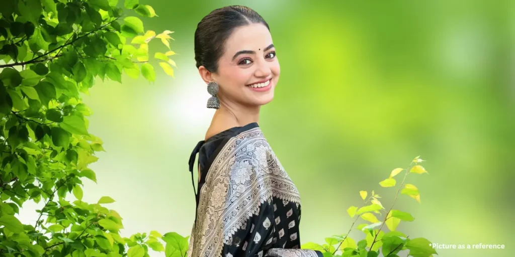 A Gujarati actress Helly Shah dressed in a black saree with intricate silver zari work, adorned with oxidized silver earrings. She is smiling brightly while posing against a vibrant green backdrop.