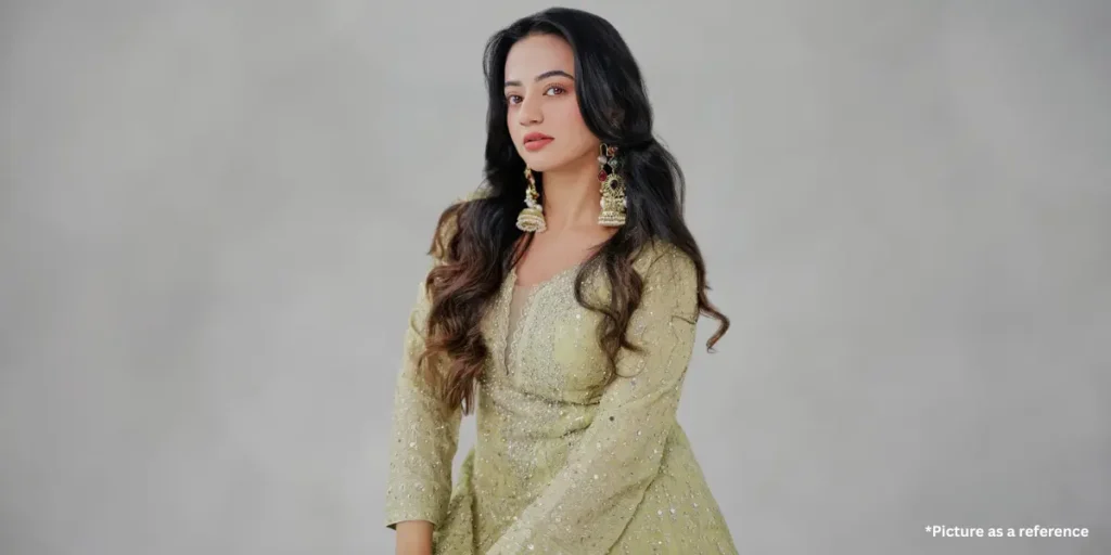 A Gujarati actress Helly Shah wearing an olive-green embroidered ethnic outfit with intricate beadwork, paired with heavy traditional earrings. She poses gracefully against a subtle grey background.