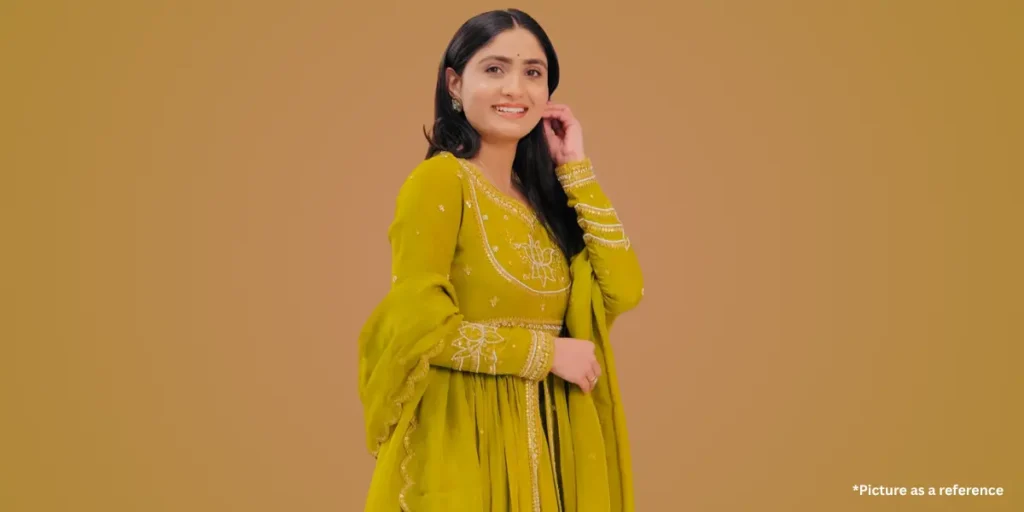 A smiling woman, Geeta Rabari, wearing a stunning mustard yellow traditional Indian dress with intricate embroidery, is posing confidently against a warm beige background.