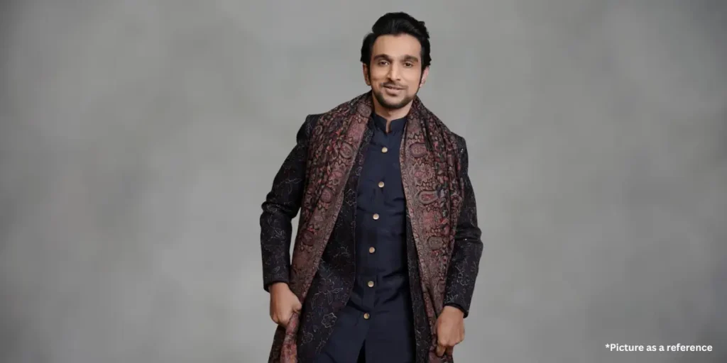 Pratik Gandhi in a traditional dark embroidered sherwani with a rich maroon shawl, standing against a grey background.