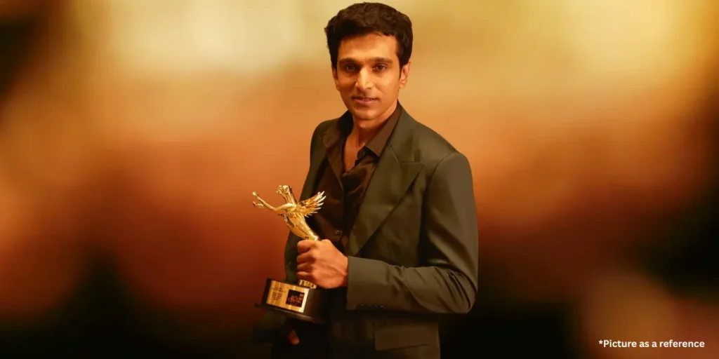Pratik Gandhi is wearing a black suit, holding a golden award trophy with a warm, glowing background.