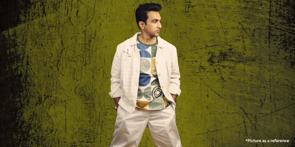 Pratik Gandhi, wearing an artistically printed T-shirt with a cream jacket and pants, poses stylishly against a distressed olive-green backdrop.