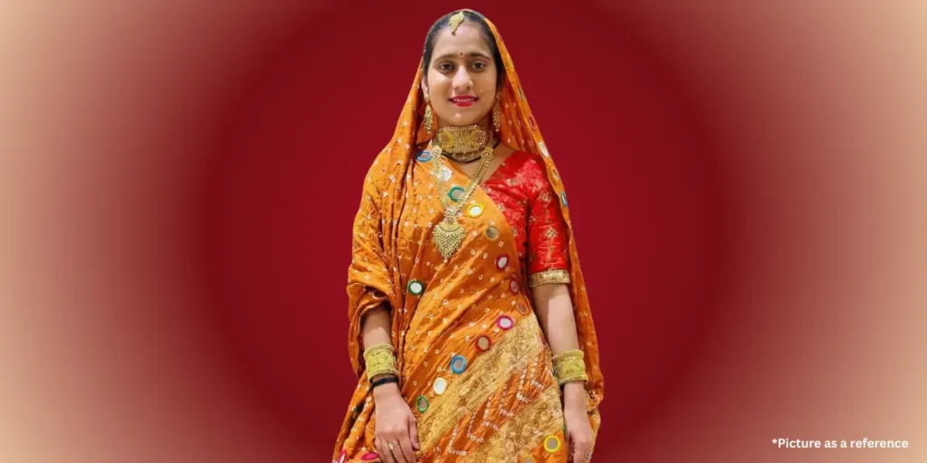 Hansha Bharvad in an elaborate orange and gold traditional Indian outfit, adorned with mirror work and a gold neckpiece and bangles.