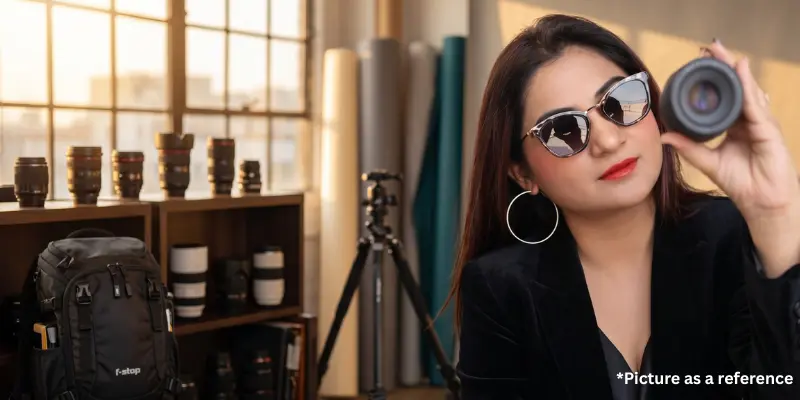 Professional woman Riya Samani wearing stylish sunglasses and hoop earrings holding a camera lens in a studio.