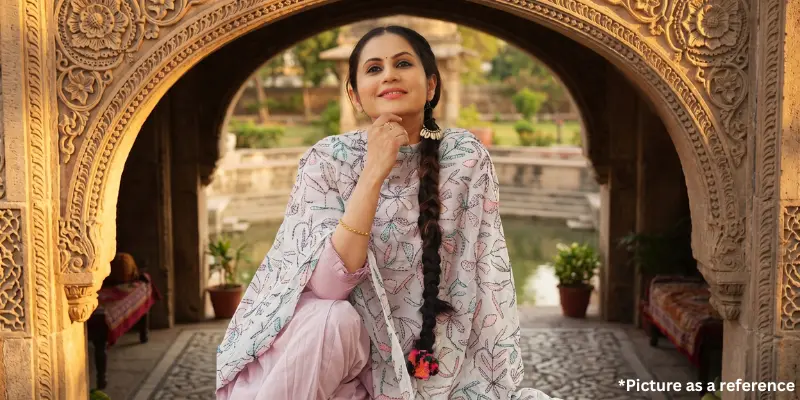 Chini Raval smiling in a light pink patterned salwar kameez with a long braid, framed by an ancient stone archway in a serene garden setting.