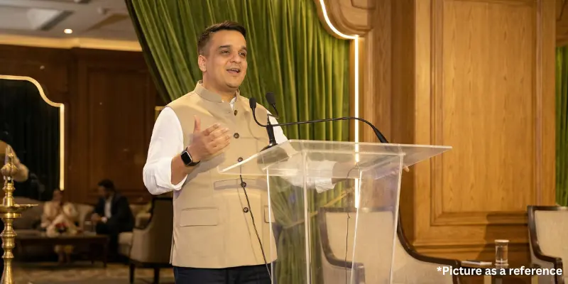 Harsh Sanghvi is giving a speech at a public event from a transparent podium, wearing a beige Nehru jacket.