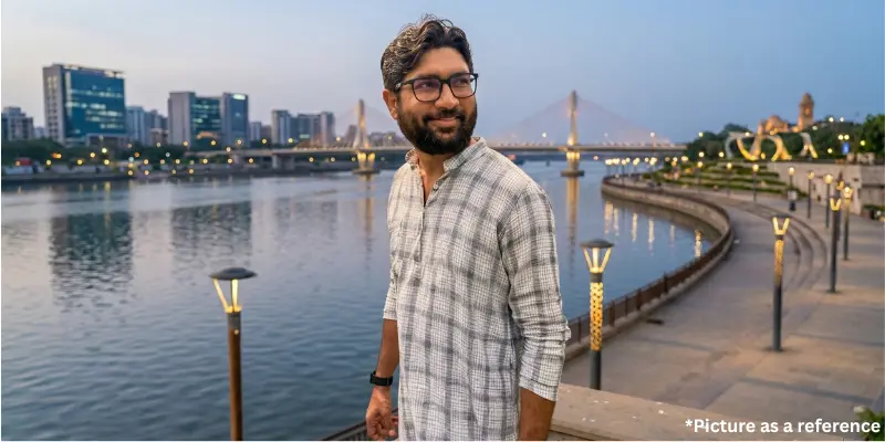 Jignesh Mevani posing at the Sabarmati Riverfront in Ahmedabad with the city skyline and bridge in the background.