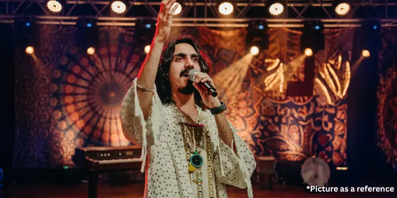 Aditya Gadhvi singing live on stage wearing a white crochet-style traditional outfit against a colorful mandala backdrop.