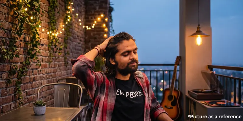 Karan Joshi running a hand through his hair with closed eyes on a brick-walled balcony featuring fairy lights, a guitar, and a vinyl record player.